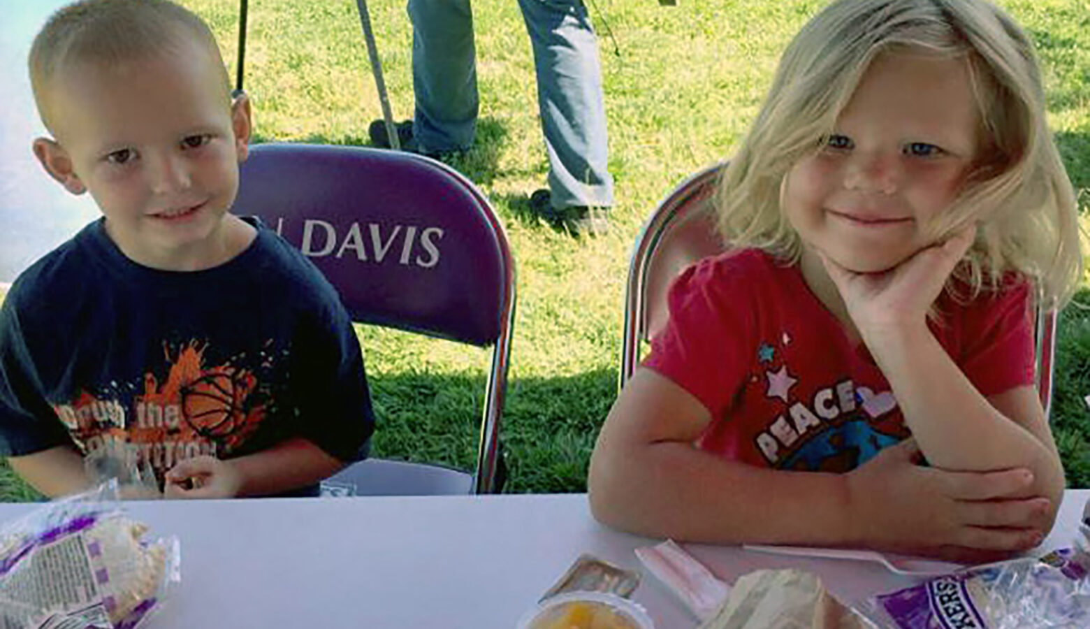Two children sit at a plastic table, posing for the camera. In front of them are lunches — Uncrustables, fruit and plasticware — provided through the Summer Food Service Program. They sit on purple folding chairs that say "BEN DAVIS."