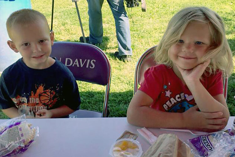 Two children sit at a plastic table, posing for the camera. In front of them are lunches — Uncrustables, fruit and plasticware — provided through the Summer Food Service Program. They sit on purple folding chairs that say "BEN DAVIS."