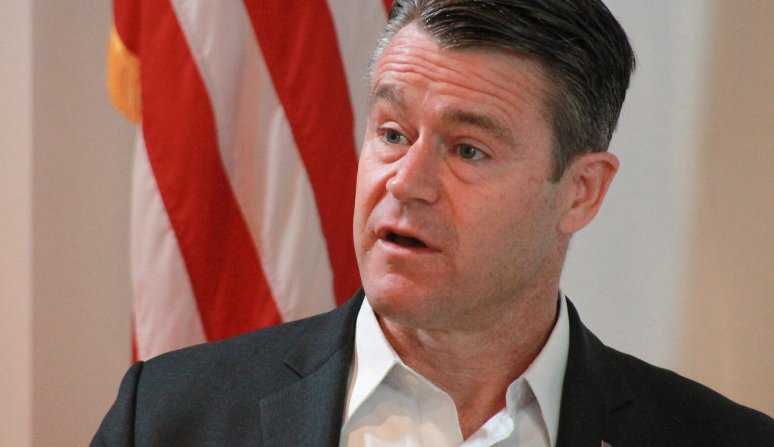 U.S. Senator Todd Young stands in front of an American flag. Young is a White man with salt and pepper hair wearing a white, collared shirt and dark gray suit jacket.
