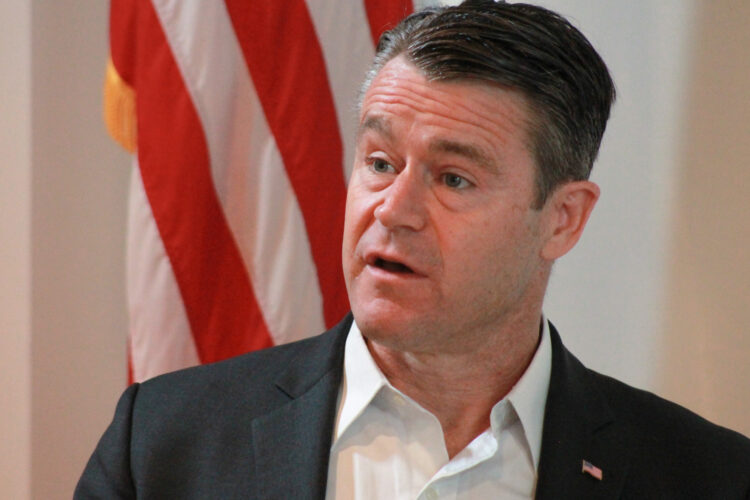 U.S. Senator Todd Young stands in front of an American flag. Young is a White man with salt and pepper hair wearing a white, collared shirt and dark gray suit jacket.