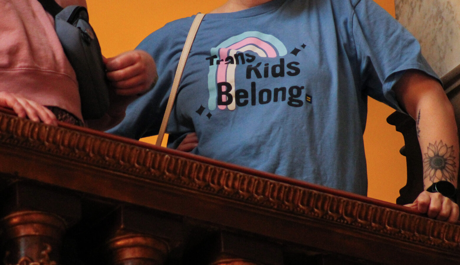A person at a protest at the Indiana Statehouse wears a t-shirt that reads, "Trans Kids Belong" with the transgender pride flag behind it.