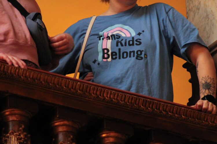 A person at a protest at the Indiana Statehouse wears a t-shirt that reads, "Trans Kids Belong" with the transgender pride flag behind it.