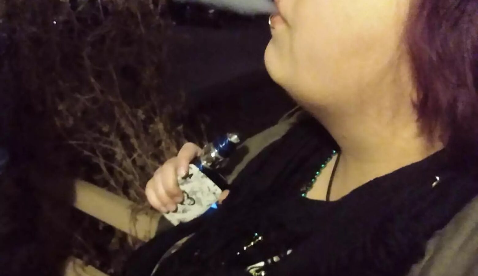 A person exhales smoke after using a vape. The person has shoulder length hair that is dyed a shade of purple and is wearing a black shirt. Only the bottom half of their face is visible in the photo.