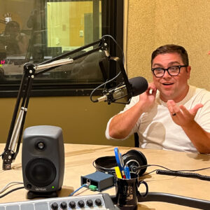 A man in a long sleeve shirt and thick glasses smiles and gestures while talking into a microphone in a radio studio.