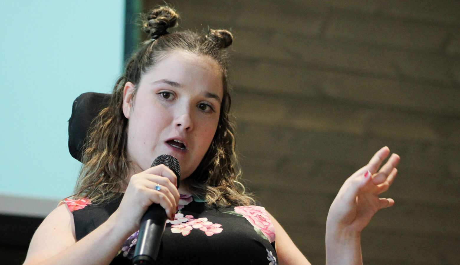 Kendra Hummer speaks into a handheld microphone at a community town hall. She is a white woman who uses a motorized wheelchair as a mobility aid. She wears a black dress with a pink flower print on it. She has brown shoulder-length hair and about half of it is in two small space buns on the top of her head. She holds the microphone in her right hand and gestures with her left hand. Behind her is the corner of a presentation screen which has a simple white slide on it.