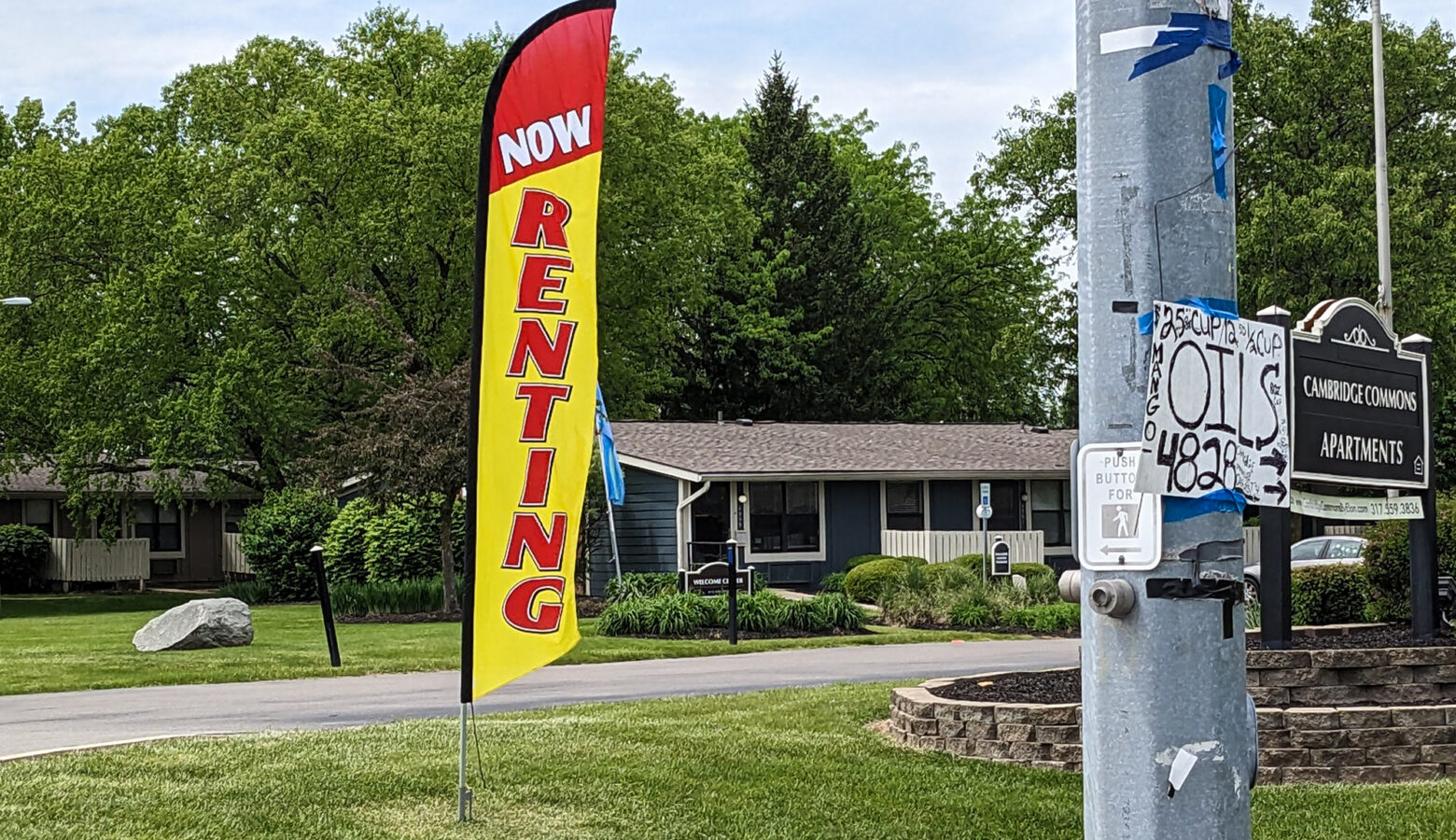 A tall sign planted in the grass outside a housing complex reads "Now Renting." The word "now" is in white letters on a red background. The word "renting" is written vertically in red letters on a yellow background.