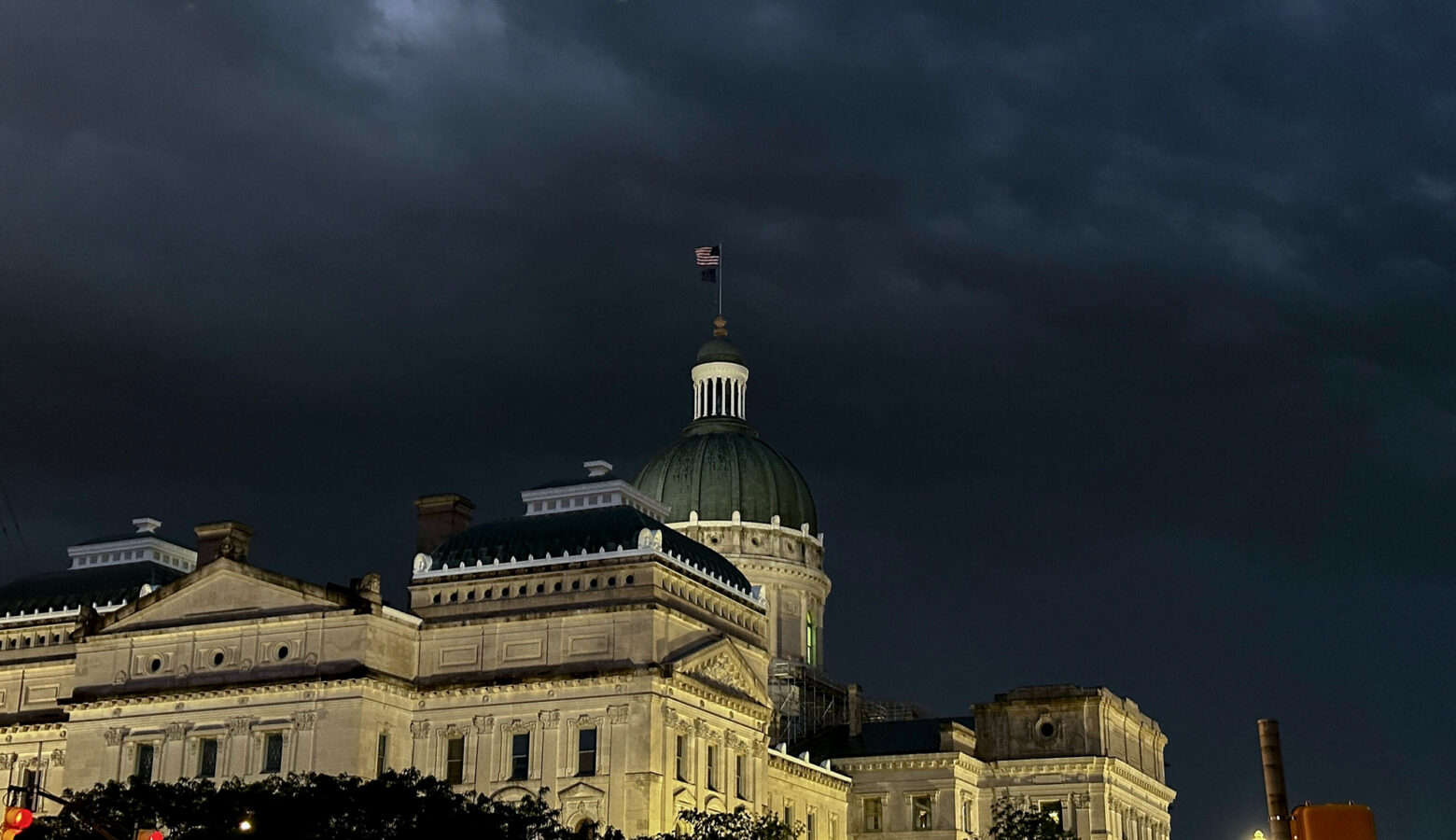 The northwestern exterior of the Indiana Statehouse is illuminated by exterior lights. Above the Statehouse are dark storm clouds. The Statehouse is a limestone building. At its center, rising above the rest of the building, is a dark green dome, topped by an American flag.