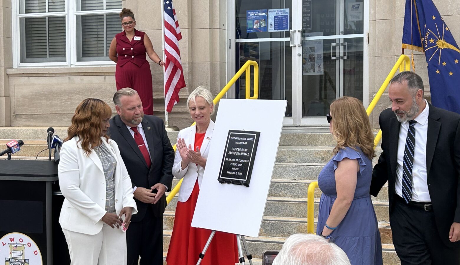 Multiple people surround a small whiteboard with a plaque hanging on it in front of the stairs to a building.