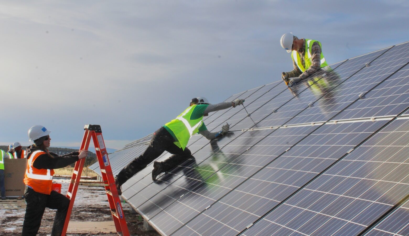 A solar installer in a bright green safety vest kneels on a solar panel while another worker in a yellow safety vest and a hard hat screws in a part. Another worker in orange holds a ladder and watches.