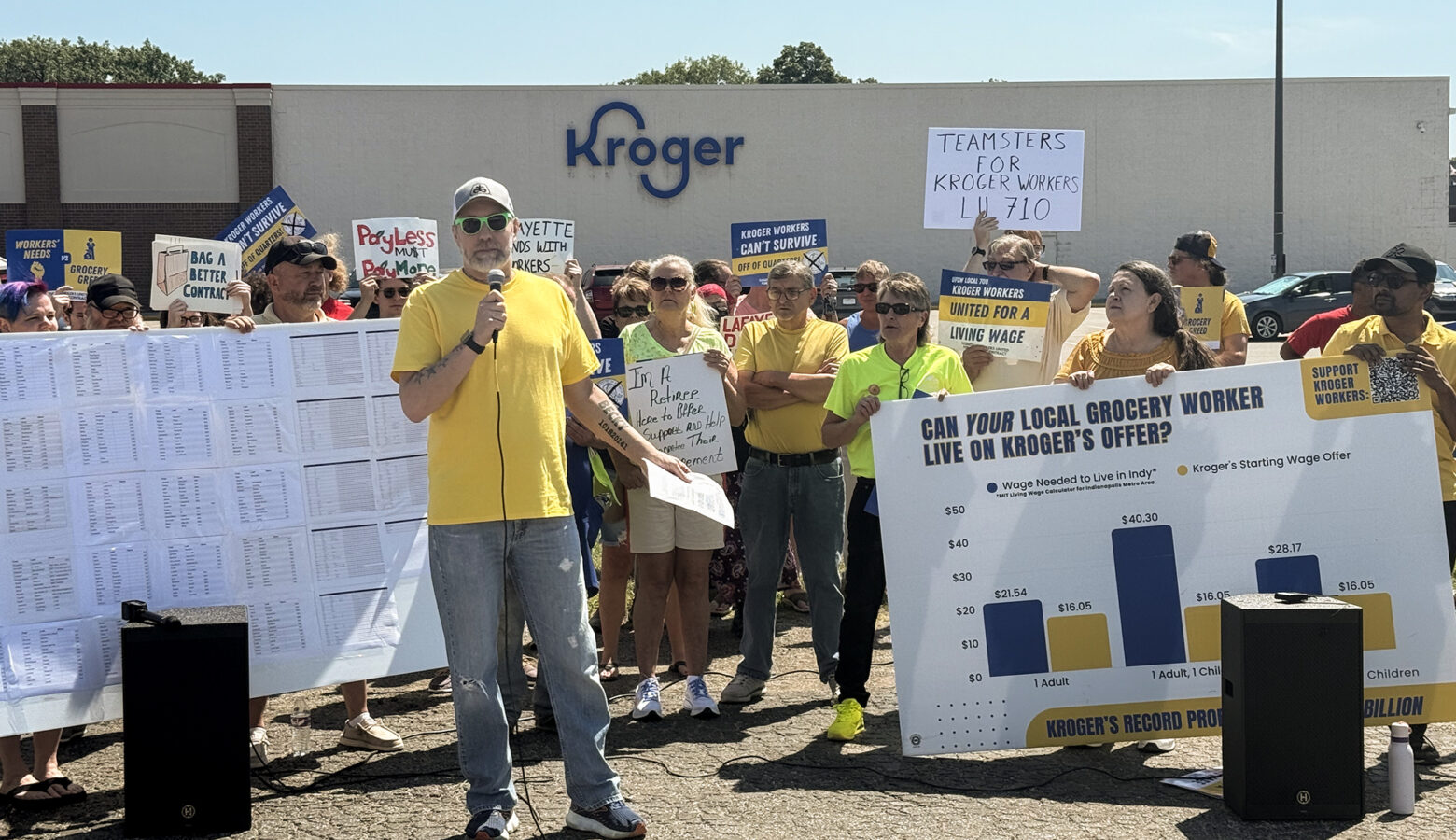 A White man speaks into a microphone wearing a bright yellow shirt and jeans with black and white shoes. A group of people stands behind him holding big white signs.
