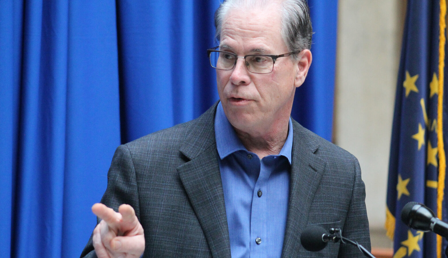 Governor Mike Braun is wearing a grey suit jacket with a blue dress shirt. He has on glasses and is looking to his right. There is a blue curtain behind him.