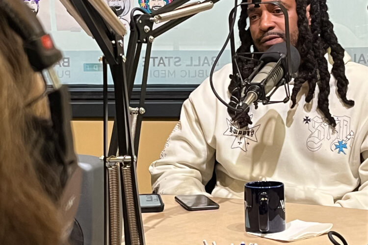 A man with long black hair and a hoodie sits behind a microphone. He is looking at a woman talking.