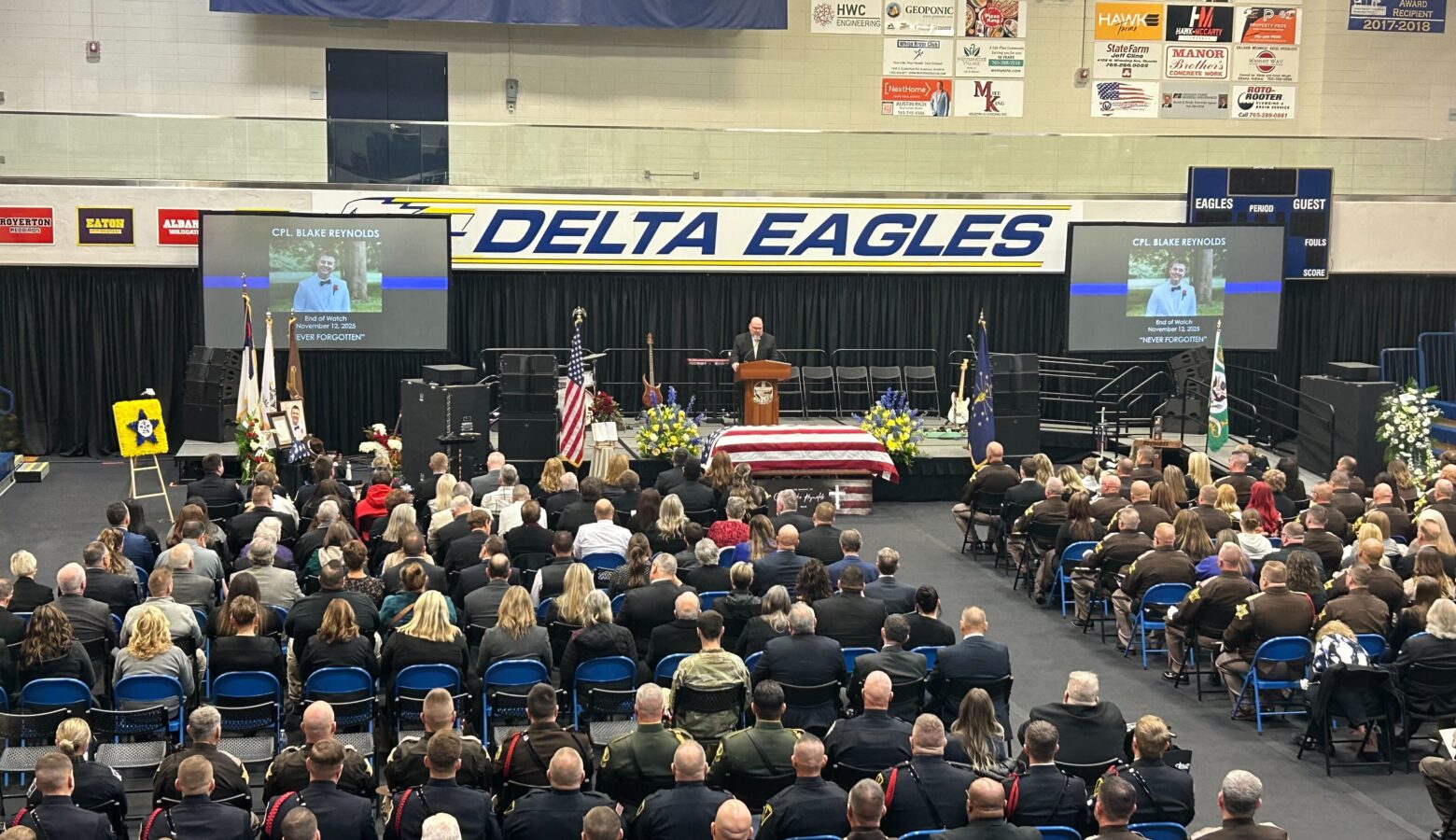 Many people sit in chairs in a high school gymnasium, listening to a speaker on a stage at the front. Two large televisions flank the stage. A banner above the stage reads Delta Eagles.