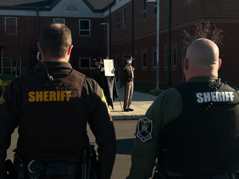 Two sheriff's deputies, seen from behind, stand in front of the Delaware County Justice Center, listening to a bell ringing.