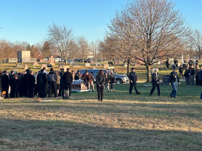 People gather around a coffin at a gravesite at a cemetery. A hearse can be seen in the back.