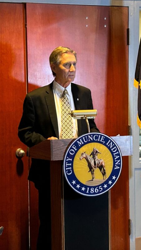 A White man stands behind a podium with a seal reading "City of Muncie Indiana." He wears a dark suit and a beige tie.