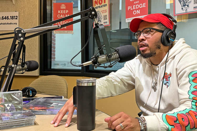 A man talks into a microphone. He has several posters, a stack of CDs, and a travel coffee mug on the table in front of him.