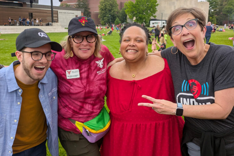 Four people in casual clothes pose for a photo smiling and laughing. There is a field with a stage behind them.
