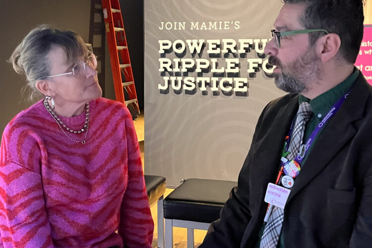 A woman and a man sit on a bench in conversation. The sign behind them has the words "powerful," "ripple," and "justice."