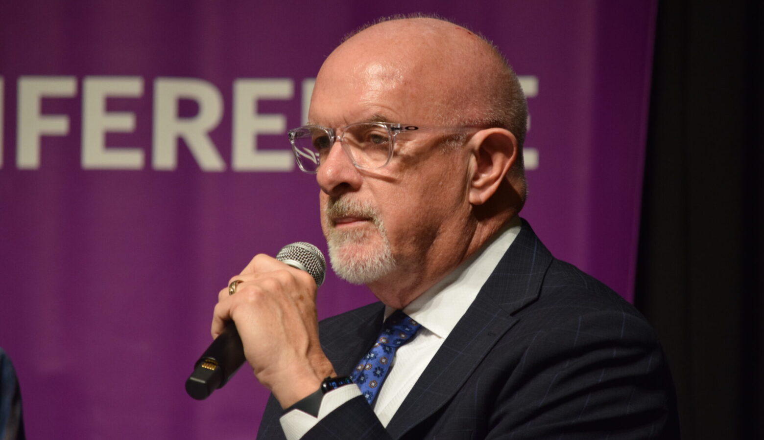 Travis Holdman holds a microphone, wearing a dark striped suit. He wears glasses.