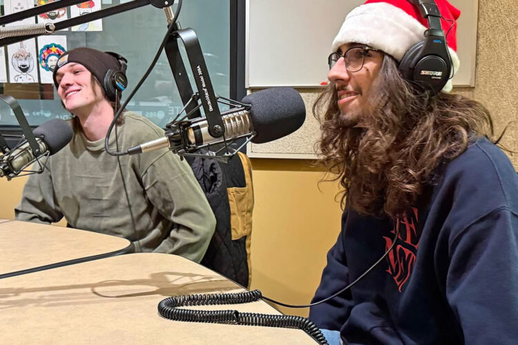 Two young men sit at a table with microphones smiling and talking. One is wearing a Santa hat.