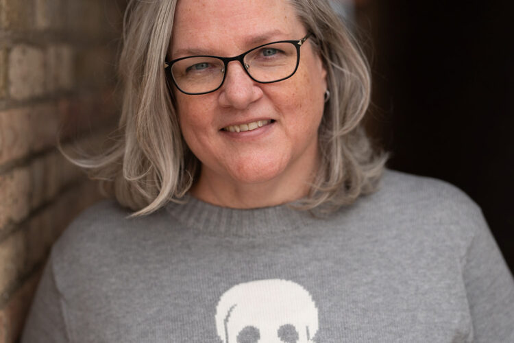 A woman smiles at the camera. She is wearing glasses and a gray sweater with a skull.