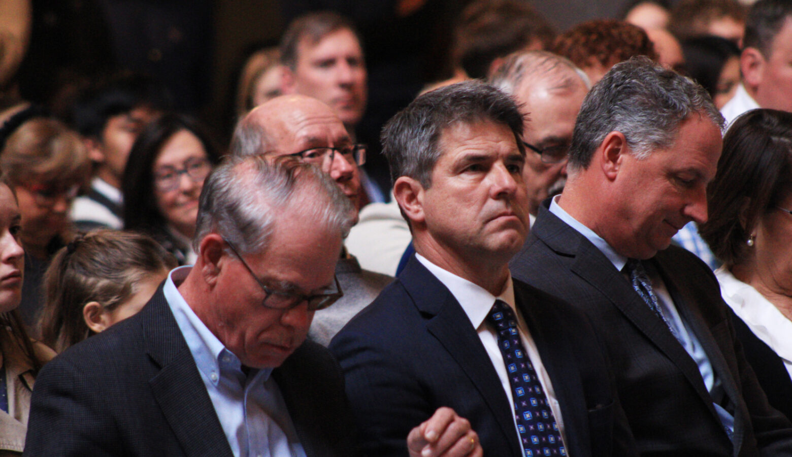 Legislative leaders sit in a crowd, listening to a speaker that is unseen.