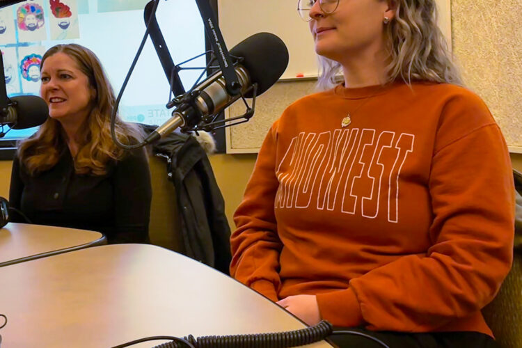 Two women sit at a table in front of microphones. They both smile at a point off-camera.