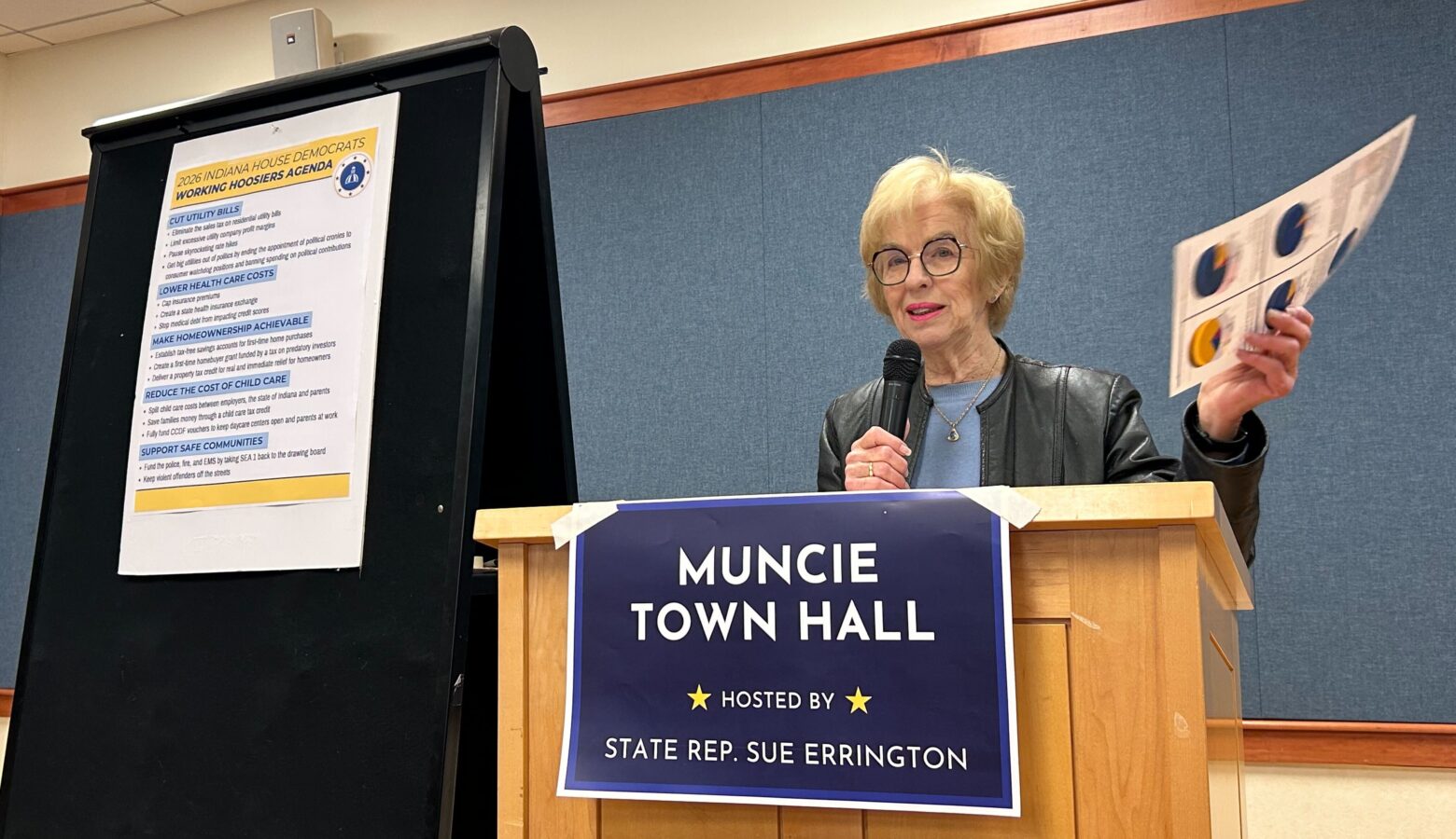 A woman stands at a podium. In her right hand she holds a microphone, in her left is a paper with statistics.