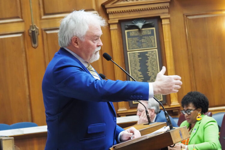 Brad Barrett is a white man with white hair and a white beard. He speaks at a podium, gesturing his hand out to the crowd.