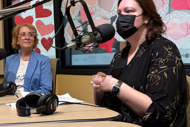Two women sit in a radio studio. One is talking and gesturing with her hands while the other listens.