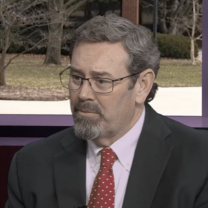 A middle-aged man with gray hair, glasses, and a neatly trimmed goatee wears a dark suit and red polka-dot tie while seated indoors, appearing thoughtful during an interview.