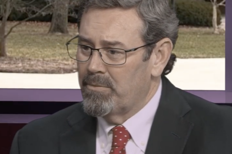 A middle-aged man with gray hair, glasses, and a neatly trimmed goatee wears a dark suit and red polka-dot tie while seated indoors, appearing thoughtful during an interview.