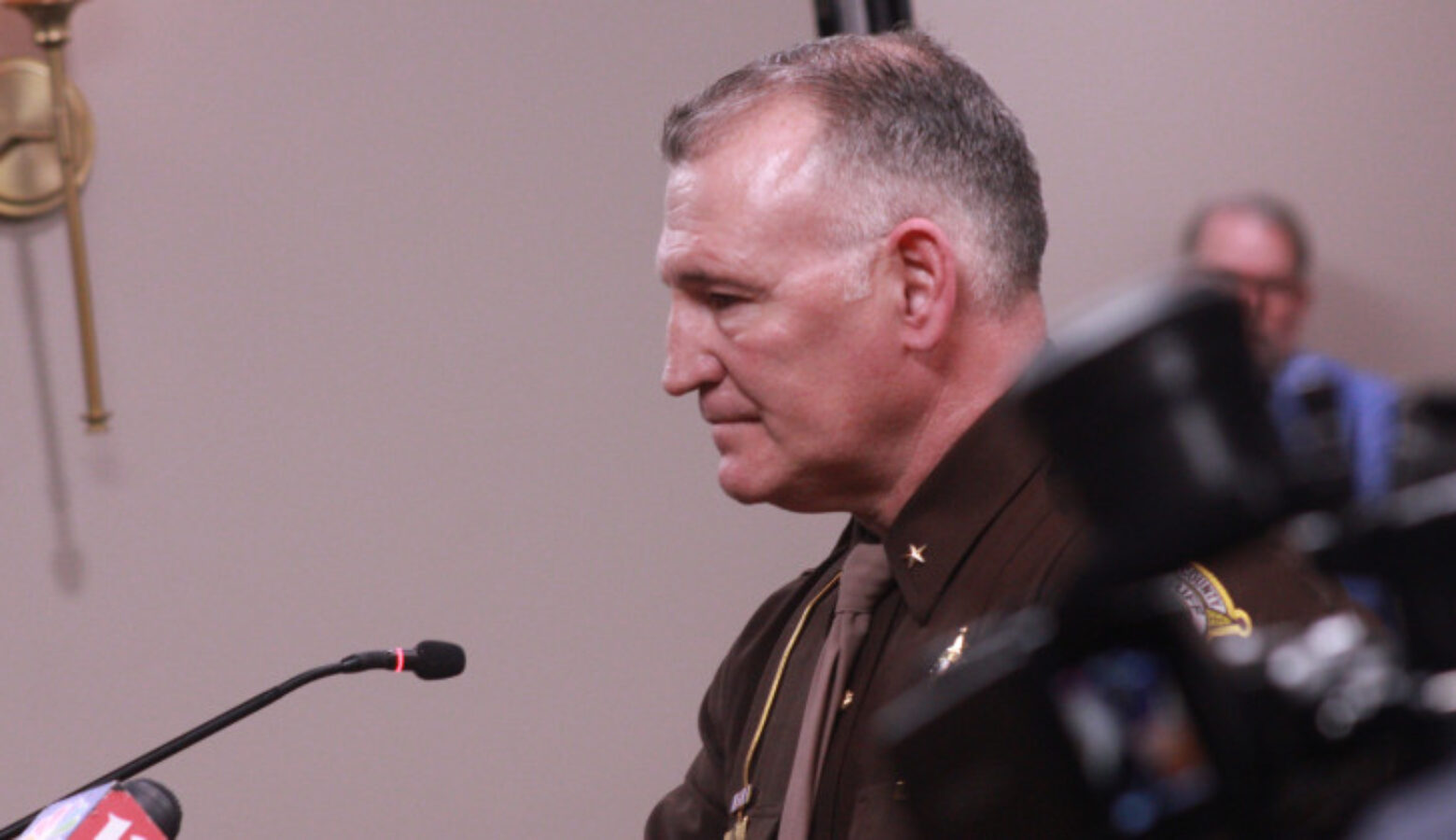 Sheriff Jeff Balon testified against the bill, arguing it would turn jails into de facto homeless shelters. He is speaking at a podium with a microphone and is photographed from the left side.