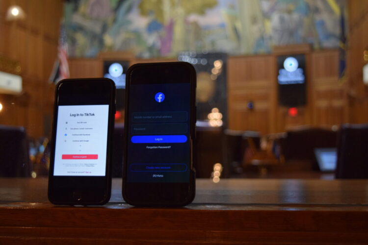 Two smartphones are tilted up displaying a Tik Tok login on one, and a Facebook login on the other. The phones are in focus in the foreground with a defocused wide shot of the Indiana House chamber in the background.