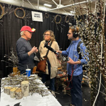 A man and a woman are talking while surrounded by willow branches made into bouquets and wreaths. Another man is holding a microphone up to them.