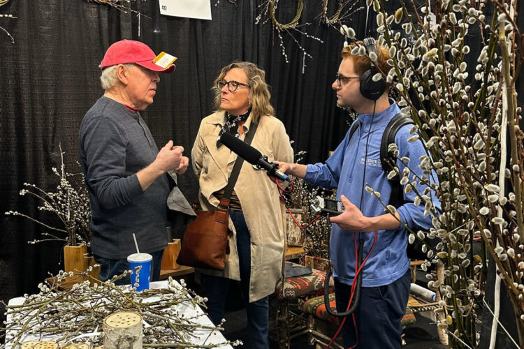 A man and a woman are talking while surrounded by willow branches made into bouquets and wreaths. Another man is holding a microphone up to them.
