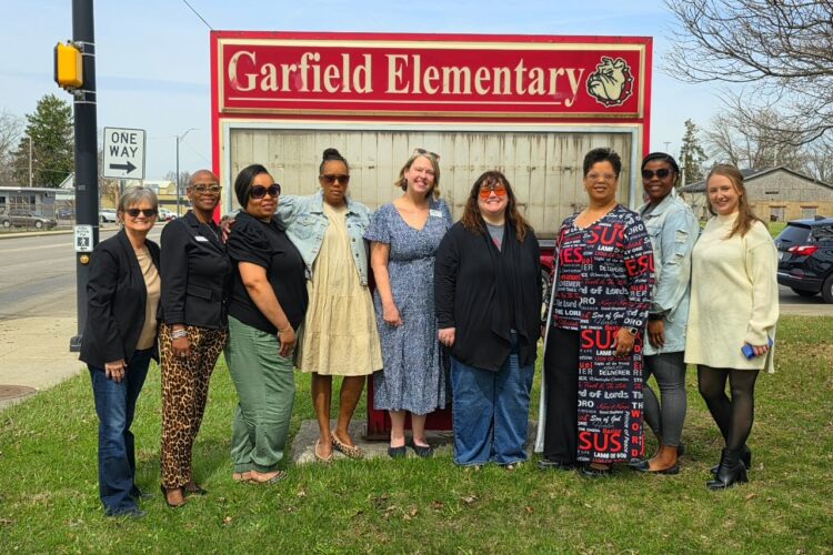 The YWCA board are nine women standing in front of a sign that reads Garfield Elementary.