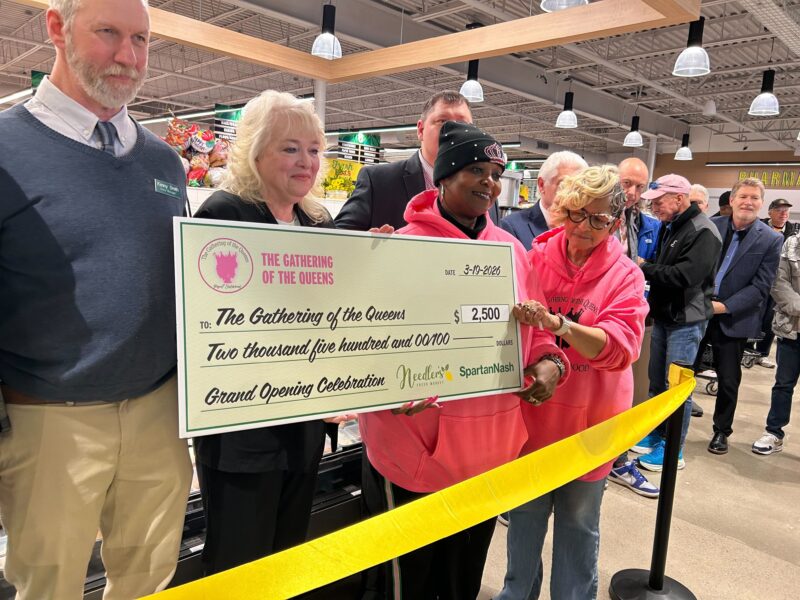 A group of people stand behind a yellow ribbon. The two women on the right where bright pink hoodies. One is holding an oversized check.