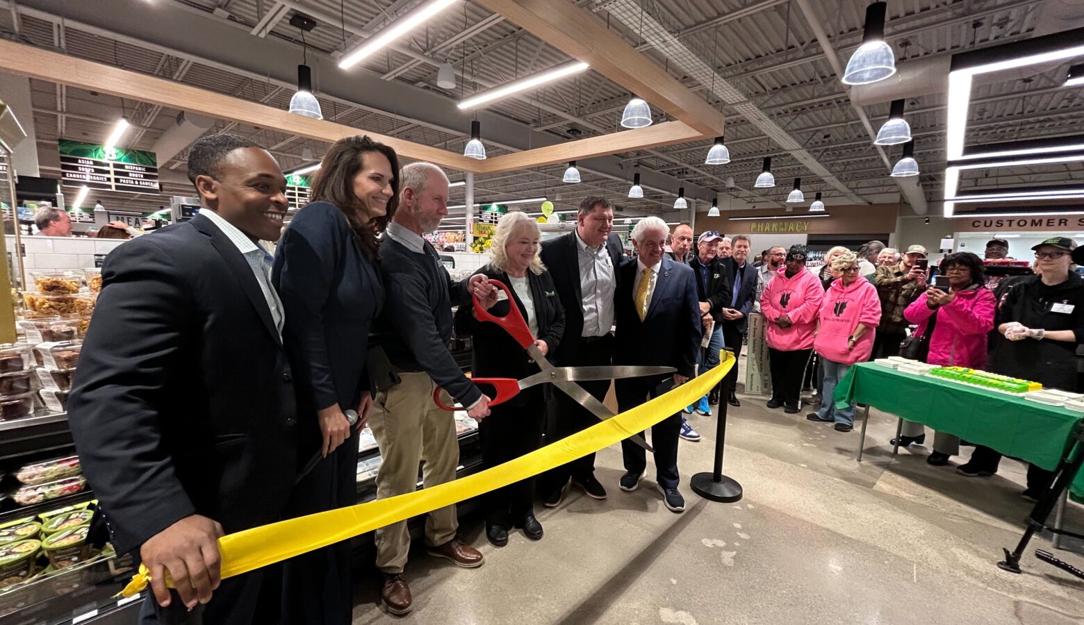 A group of people stand behind a yellow ribbon. A man in the middle of them holds a large pair of scissors preparing to cut the ribbon