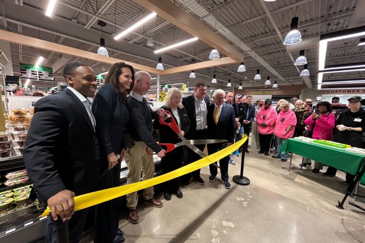 A group of people stand behind a yellow ribbon. A man in the middle of them holds a large pair of scissors preparing to cut the ribbon