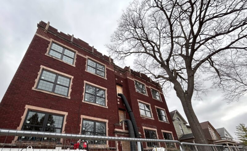 The side of a large brick building with a large air tube going into the buildings third floor. A large tree sits just to the right of it.