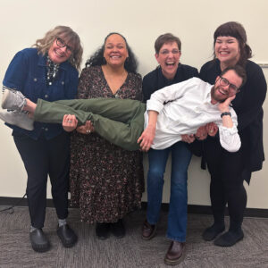 Four women are holding up a man horizontally across all of them. They are all smiling and laughing.