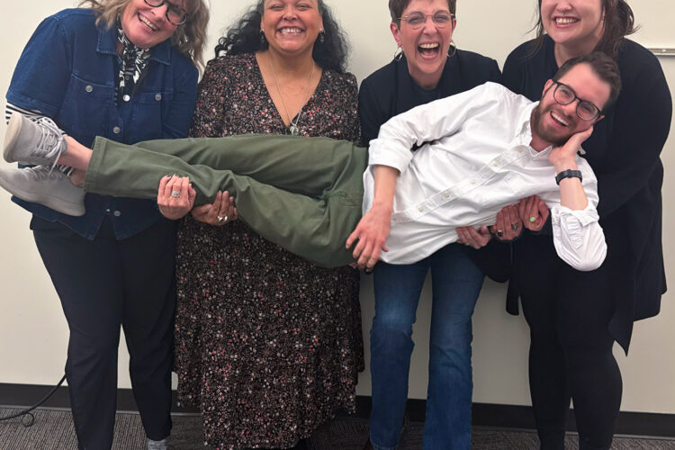 Four women are holding up a man horizontally across all of them. They are all smiling and laughing.