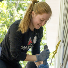 A young woman with a ponytail smiles while using a hand tool during a home-building project, wearing work gloves and a Ball State softball shirt.