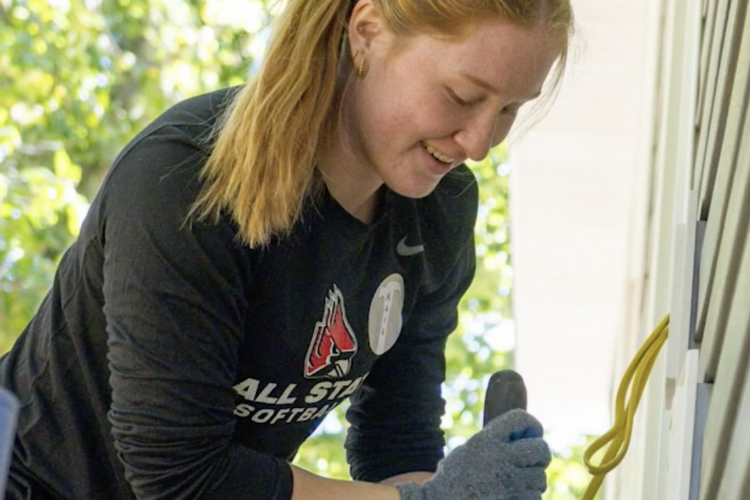 A young woman with a ponytail smiles while using a hand tool during a home-building project, wearing work gloves and a Ball State softball shirt.