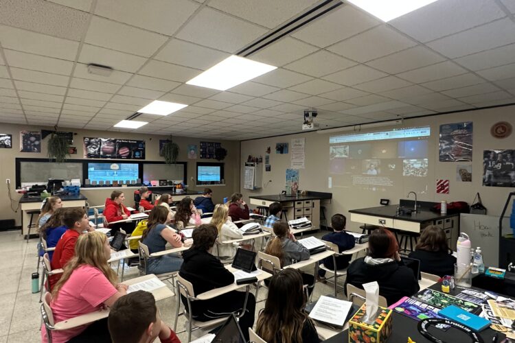 A class of students sit in desks and face a projection of an online meeting on the white board.
