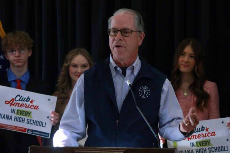 Governor Mike Braun stands in front of a podium, speaking into a microphone. Behind him, high school students stand listening, holding up signs that say "Club America in every Indiana high school."