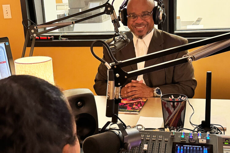 A man is sitting in a small radio studio wearing headphones. He smiles while listening.