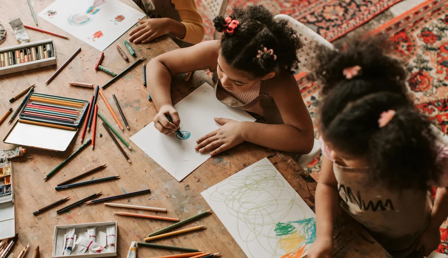 Two girls are pictured from above using crayons and pencils and paper on a table to make drawings.